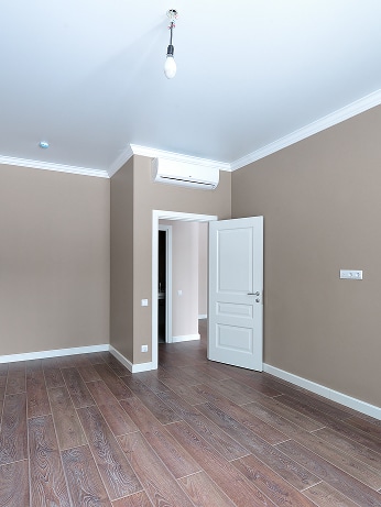 Pièce vide appartement neuf avec murs beiges, parquet au sol, climatisation, porte blanche ouverte et ampoule au plafond.