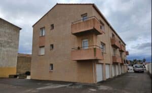Immeuble résidentiel de plusieurs étages avec façades claires, balcons orangés et garages en rez-de-chaussée. Ciel nuageux.