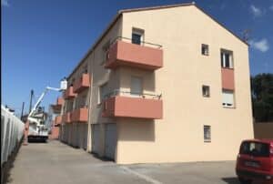 Bâtiment beige balcons saumon, ouvrier en nacelle rénove la façade près du toit. Garages et voiture rouge (FR-836-JD).